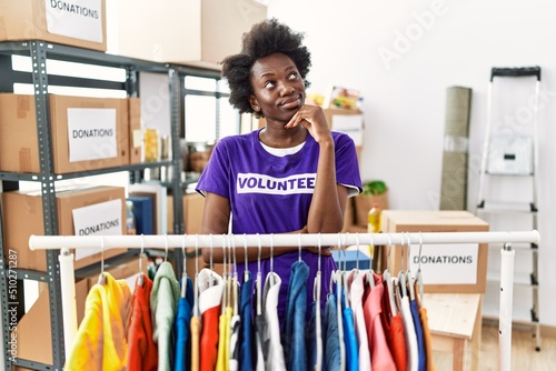 African young woman wearing volunteer t shirt at donations stand with hand on chin thinking about question, pensive expression. smiling with thoughtful face. doubt concept.