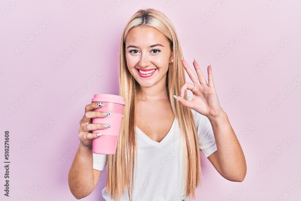 Young blonde girl holding coffee doing ok sign with fingers, smiling friendly gesturing excellent symbol Young blonde girl holding coffee doing ok sign with fingers, smiling friendly gesturing excellent symbol