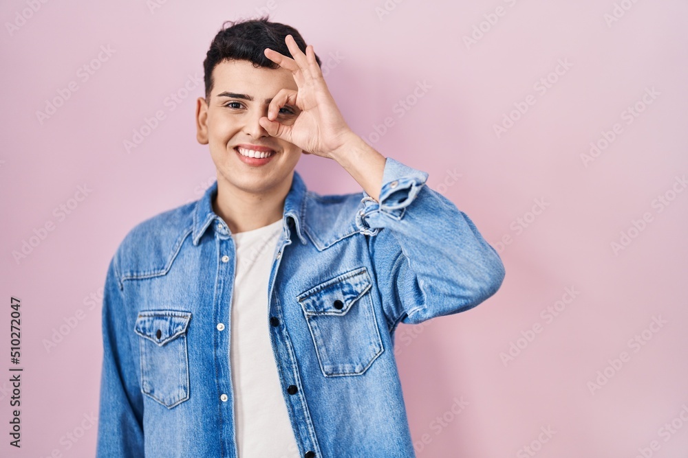 Non binary person standing over pink background smiling happy doing ok sign with hand on eye looking through fingers Non binary person standing over pink background smiling happy doing ok sign with hand on eye looking through fingers