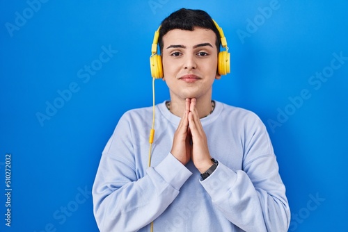 Non binary person listening to music using headphones praying with hands together asking for forgiveness smiling confident.