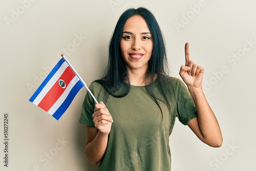 Young hispanic girl holding costa rica flag smiling with an idea or question pointing finger with happy face, number one