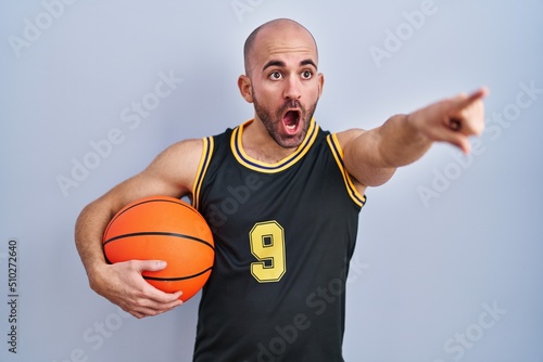 Young bald man with beard wearing basketball uniform holding ball pointing with finger surprised ahead, open mouth amazed expression, something on the front