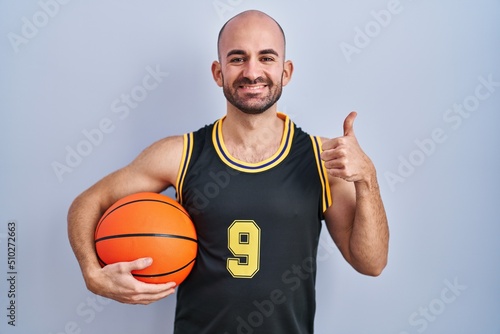 Young bald man with beard wearing basketball uniform holding ball doing happy thumbs up gesture with hand. approving expression looking at the camera showing success.