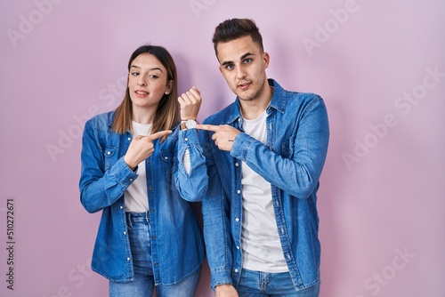 Young hispanic couple standing over pink background in hurry pointing to watch time, impatience, looking at the camera with relaxed expression