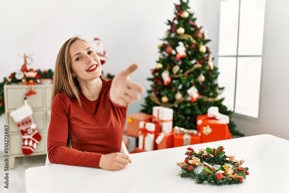 Caucasian young blonde woman sitting on the table by christmas tree smiling friendly offering handshake as greeting and welcoming. successful business. Caucasian young blonde woman sitting on the table by christmas tree smiling friendly offering handshake as greeting and welcoming. successful business.