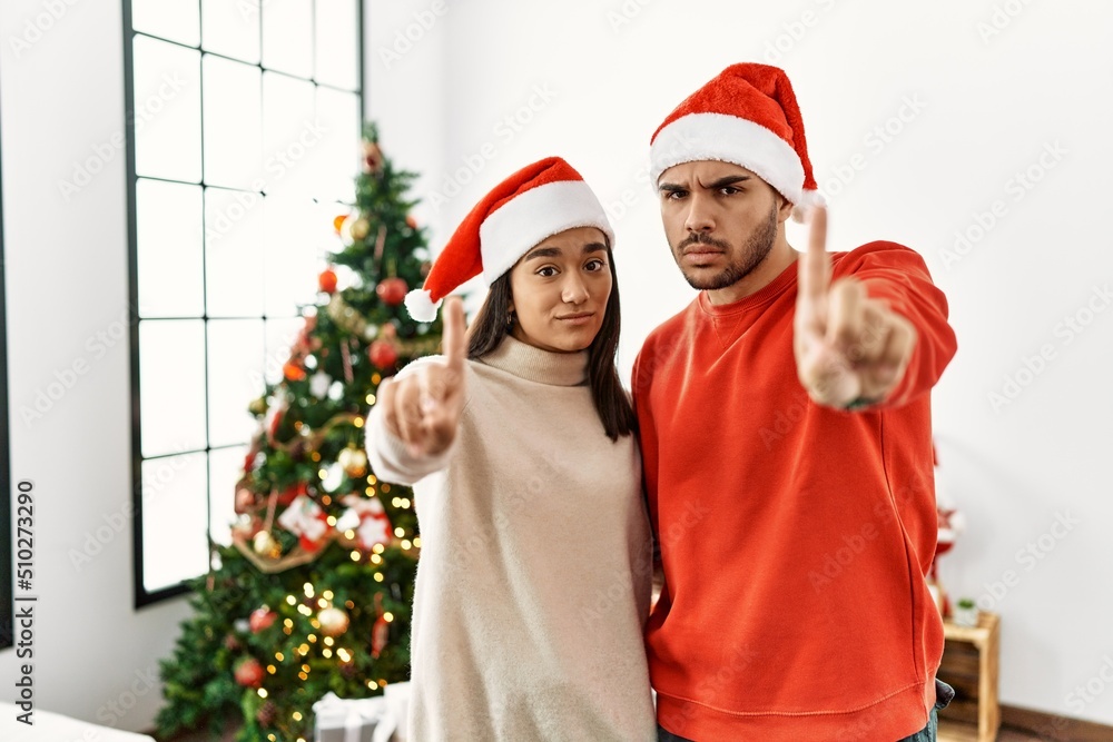 Young hispanic couple standing by christmas tree pointing with finger up and angry expression, showing no gesture Young hispanic couple standing by christmas tree pointing with finger up and angry expression, showing no gesture
