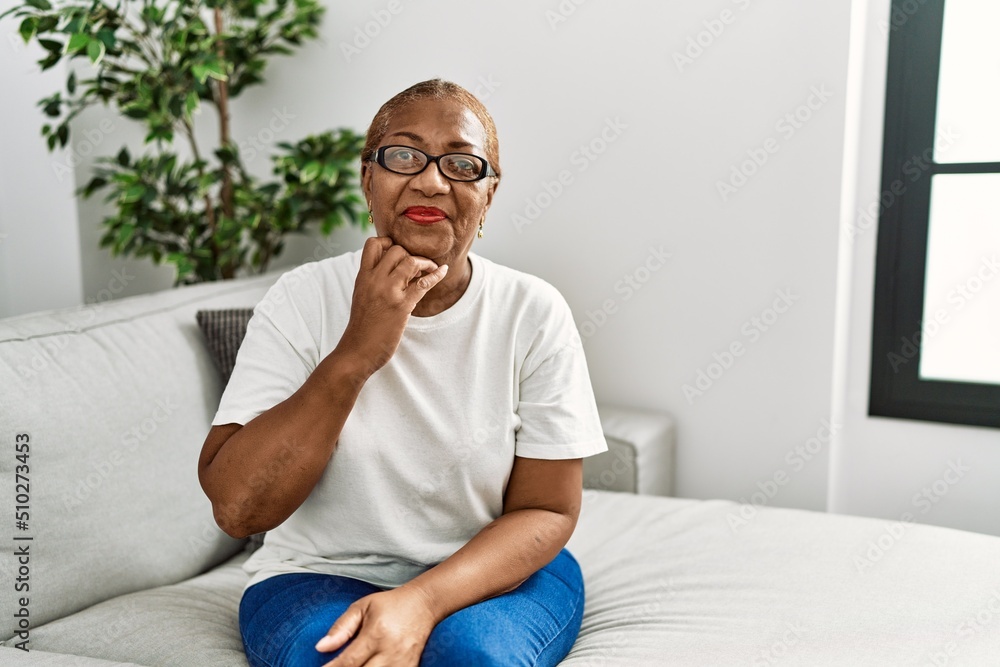 Mature hispanic woman sitting on the sofa at home with hand on chin thinking about question, pensive expression. smiling with thoughtful face. doubt concept.