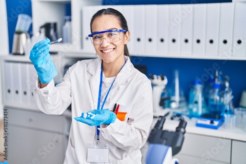 Young hispanic woman scientist holding pills with tweezer at laboratory