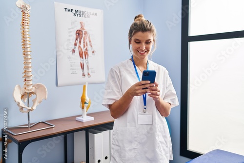 Young blonde woman wearing physiotherapist uniform using smartphone at clinic