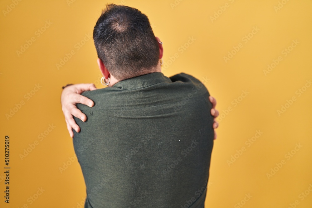 Plus size hispanic man with beard standing over yellow background hugging oneself happy and positive from backwards. self love and self care