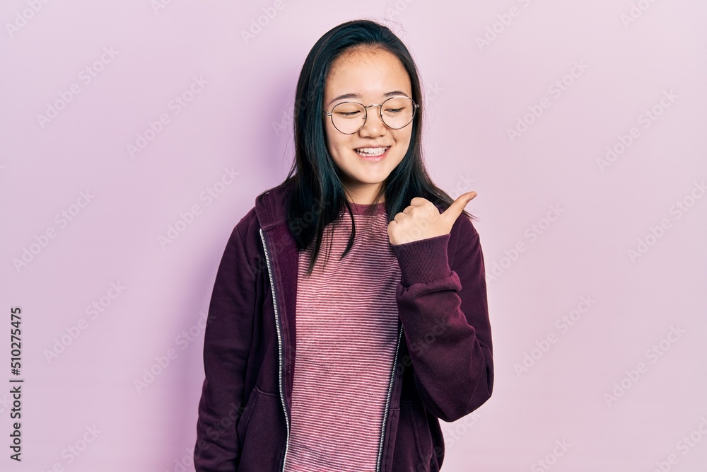 Young chinese girl wearing casual clothes and glasses smiling with happy face looking and pointing to the side with thumb up. Young chinese girl wearing casual clothes and glasses smiling with happy face looking and pointing to the side with thumb up.