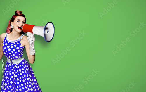 Portrait image of purple haired woman holding mega phone, shout advertising something. Girl in blue pin up style dress with mega phone loudspeaker. Isolated on green background. Beauty model.