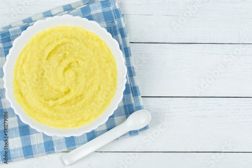 Corn porridge for a baby made of ground cereals in a white bowl with a banana on a blue napkin on a white wooden background. Space for text. Baby's first complementary food, baby nutrition.