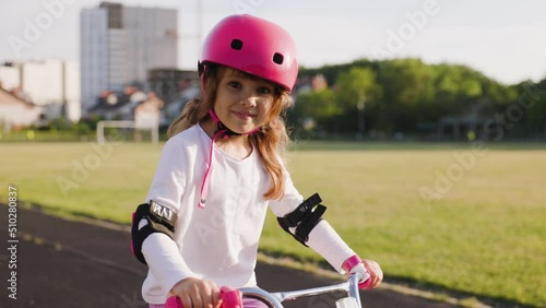 Cute light hair little girl in pink helmet in elbow and knee pads rides a bicycle at the stadium. People playing football in background. Sport activity for children. dolly-in shot photo