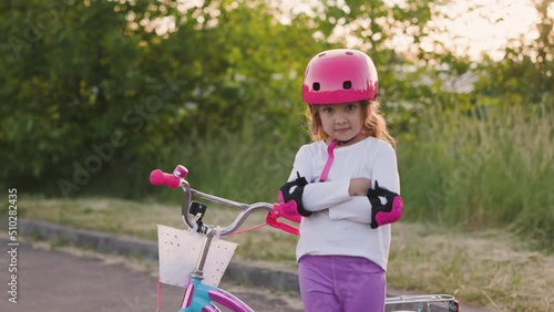 Cute light hair little girl in pink helmet in elbow and knee pads folds her hands next to bicycle in the park. Sport activity for children photo