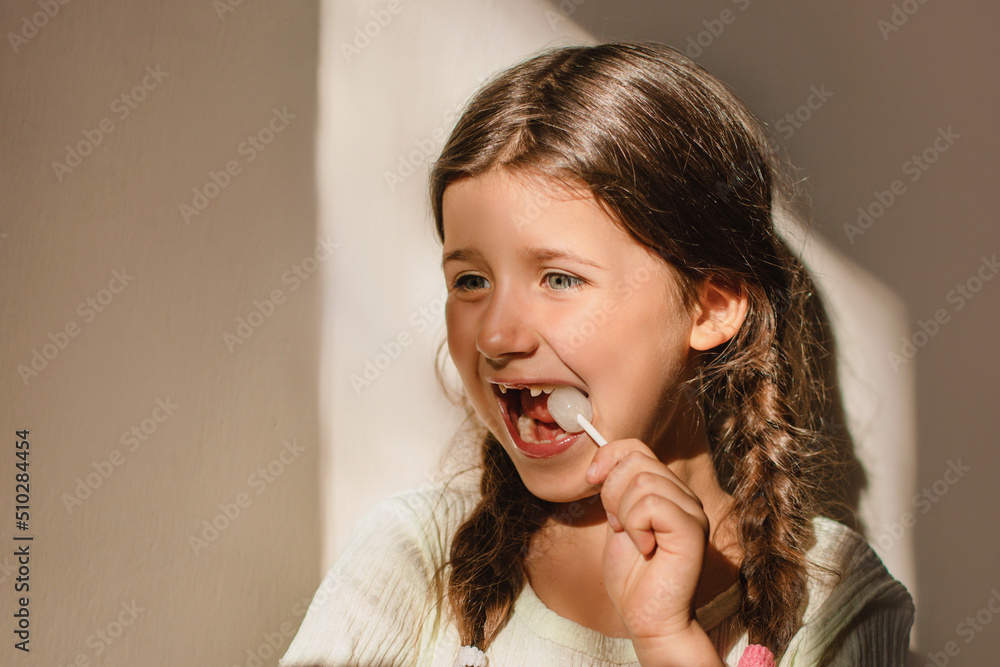 Child girl joyfully eats a lollipop indoors in the sun. baby teeth health concept Child girl joyfully eats a lollipop indoors in the sun. baby teeth health concept