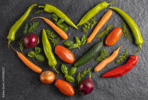 Heart made of fresh vegetables: green pepper, tomato, onion, carrot, cucumber, basil, dill on grey black background