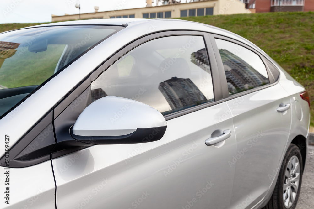 Close-up of the side right mirror and window of the car body silver sedan on the street parking after washing and detailing in auto service industry. Road safety while driving.