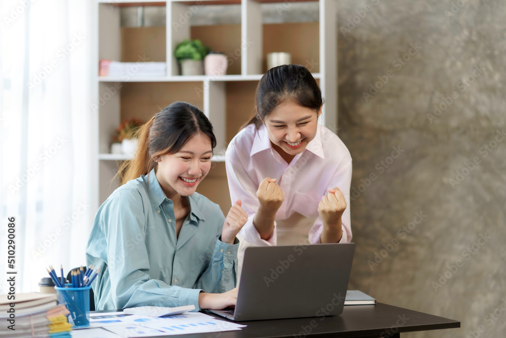 Two young Asian businesswomen show joyful expression of success at work smiling happily with a laptop computer in a modern office. Two young Asian businesswomen show joyful expression of success at work smiling happily with a laptop computer in a modern office.