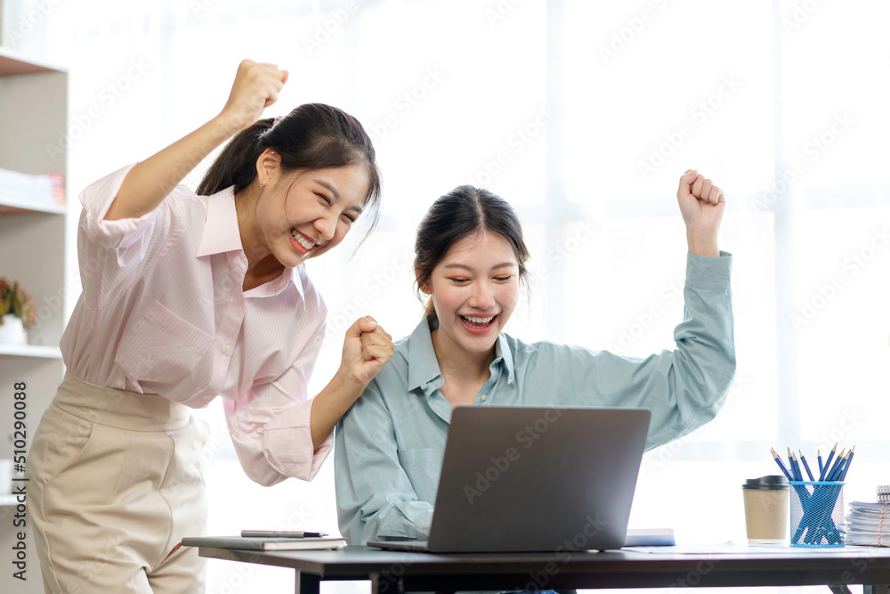 Two young Asian businesswomen show joyful expression of success at work smiling happily with a laptop computer in a modern office. Two young Asian businesswomen show joyful expression of success at work smiling happily with a laptop computer in a modern office.