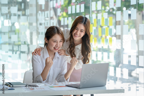 Two young Asian businesswomen show joyful expression of success at work smiling happily with a laptop computer in a modern office.