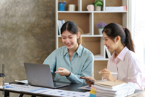 Two young beautiful Asian business woman in the conversation, exchanging ideas on laptop computer together at office.