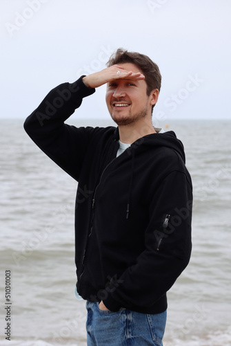 A young man in a black hoodie on the beach against the backdrop of the sea. Brown hair, blue eyes, smiling © Andrei Antipov