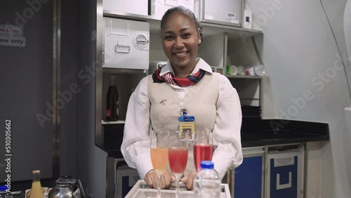 Portrait of happy African woman flight attendant in uniform standing and looking camera at kitchen room in airplane. Attractive young female stewardess smiling and serving food and drink to passenger. photo