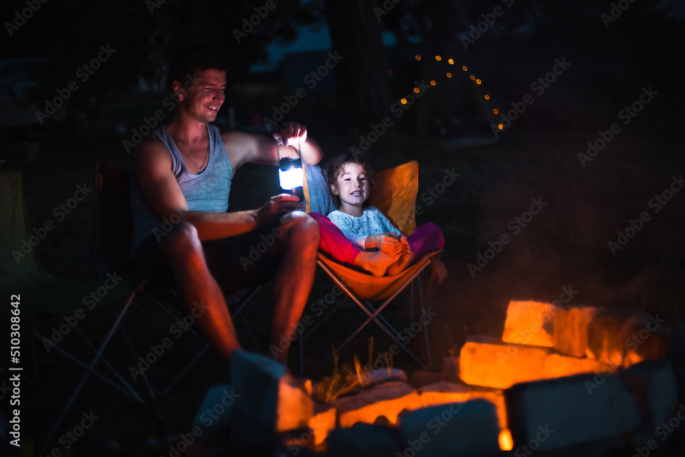 Dad and daughter sit at night by the fire in the open air in the summer in nature. Family camping trip, gatherings around the campfire. Father's Day, barbecue. Camping lantern and tent