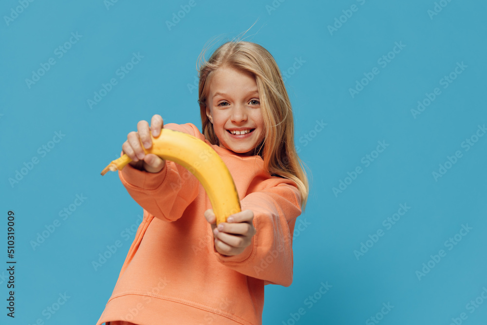 a cute, happy school-age girl stands on a blue background in bright clothes and holds a banana in her hand a cute, happy school-age girl stands on a blue background in bright clothes and holds a banana in her hand