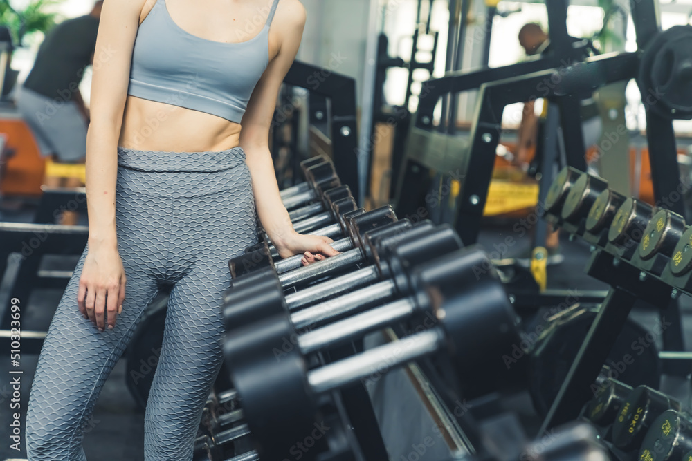 Fototapeta premium Unrecognizable caucasian slim woman wearing sports bra and patterned leggings, reaching for a big dumbbell. Indoor shot. High quality photo