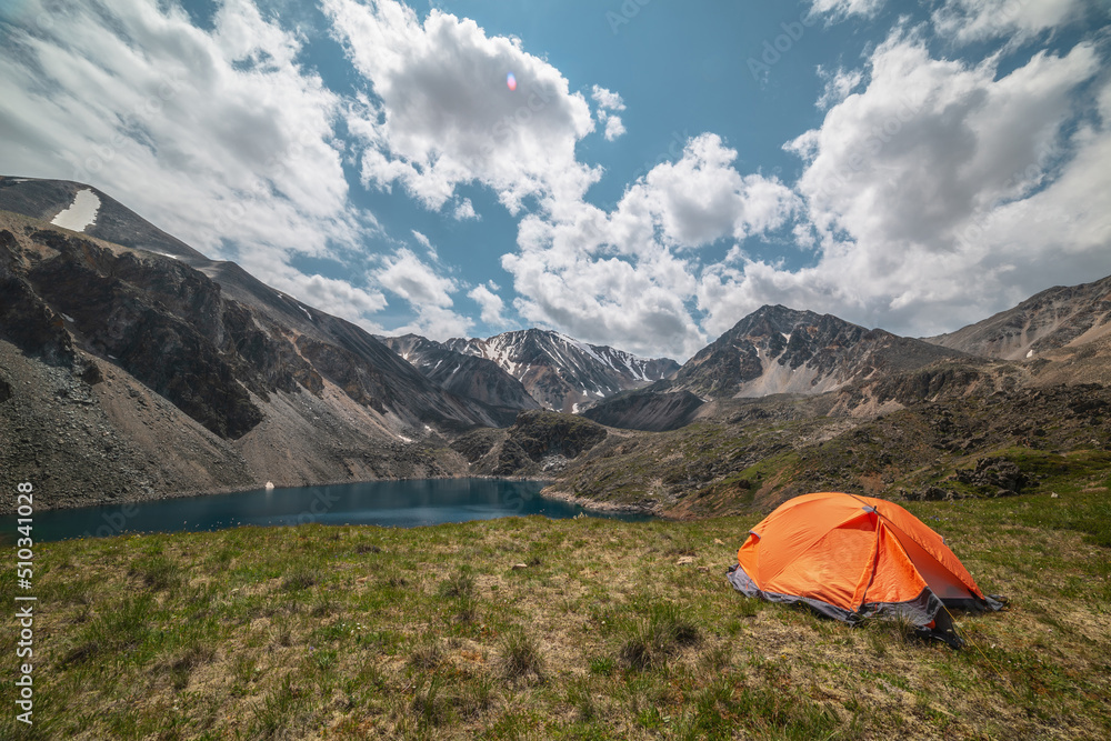 Vivid orange tent on grassy hill with view to mountain lake of phantom blue color among high mountains in changeable weather. Tent near deep blue lake among sunlit large mountains under cloudy sky.