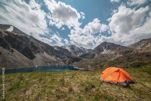 Vivid orange tent on grassy hill with view to mountain lake of phantom blue color among high mountains in changeable weather. Tent near deep blue lake among sunlit large mountains under cloudy sky. © Daniil