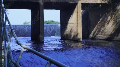Camera zoom in forward. Shade and view through channel of watercourse under the bridge. Old concrete dam with water flowing rapids. During the rainy season in rural Thailand.