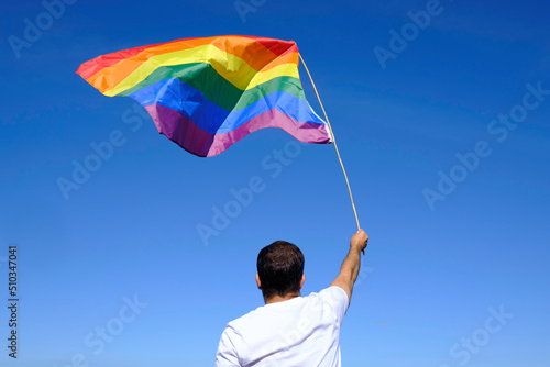 Adult man in white t-shirt waves rainbow flag with blue sky and ocean in the background.