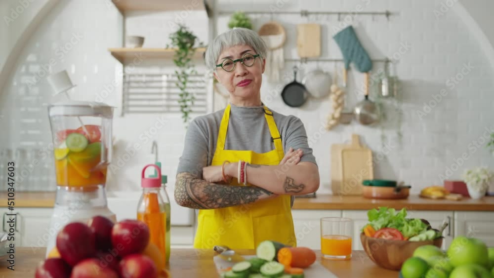 Mature smiling tattoo woman eating salad, fruits and vegetables. Attractive mature woman with fresh green fruit salad at home. Senior woman apron standing in the kitchen counter relaxing in house Mature smiling tattoo woman eating salad, fruits and vegetables. Attractive mature woman with fresh green fruit salad at home. Senior woman apron standing in the kitchen counter relaxing in house