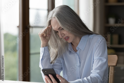 Angry nervous older 50s businesswoman sit at desk look at smartphone screen, feel stressed read sms with bad news, dissatisfied with broken device, missed call, professional or gadget problems concept