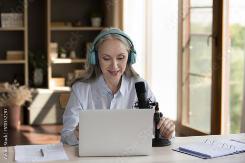 Middle-aged attractive woman wear headphones speaks into professional microphone sits at desk with laptop. Live stream event, radio host profession, speaker leads program on air, broadcasting concept