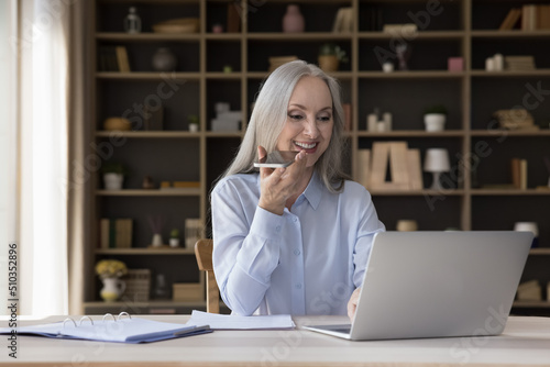 Middle-aged businesslady holds smartphone talks on speakerphone sit at desk, leave voice message via messenger, text on laptop. Older generation use modern tech for business and communication concept