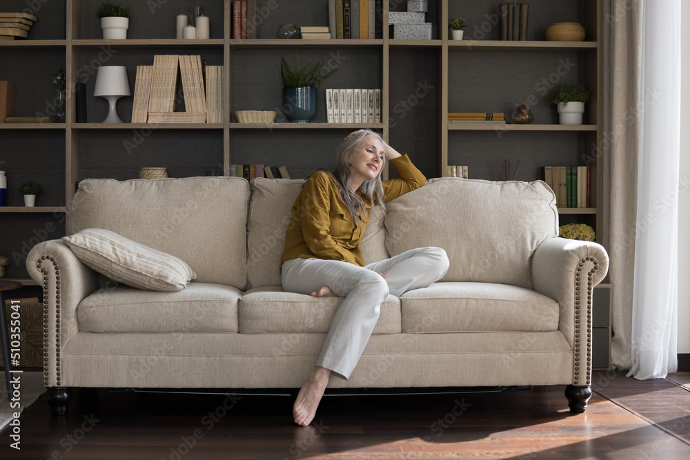 In cozy light living room middle-aged serene beautiful woman sit on sofa looks pensive enjoy free time, daydreaming relaxing alone at home. Homeowner person, tenancy, modern furniture store ad concept