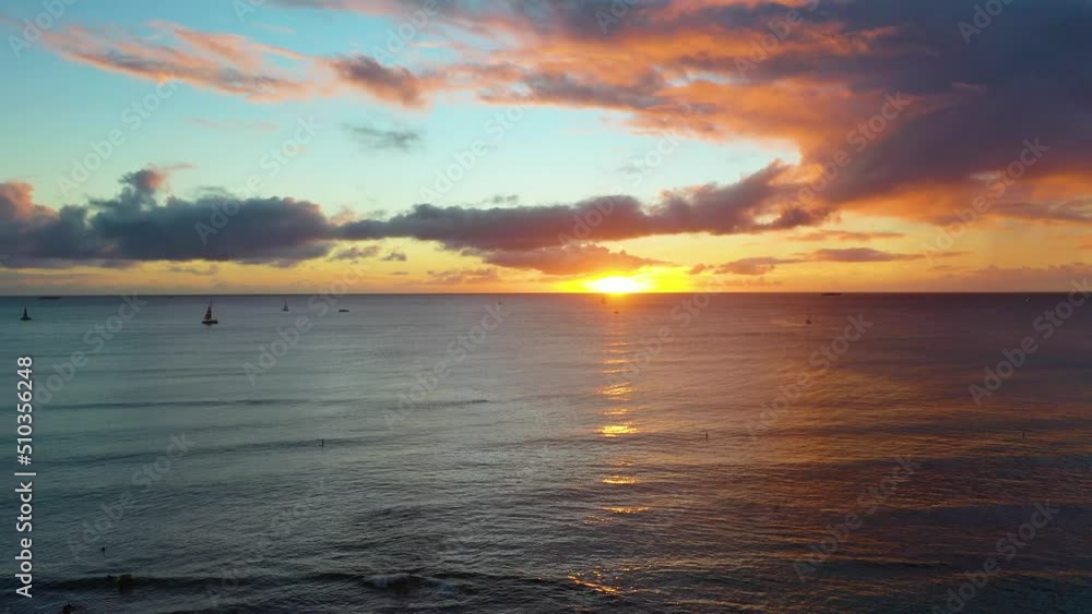 Sun Setting over Sailboats And Surfers in the Rippling Waves of Waikiki Beach in Honolulu, Hawaii. Reflecting Sunlight as they Sail and Swim in the Ocean. Aerial Drone Pan Left Sun Setting over Sailboats And Surfers in the Rippling Waves of Waikiki Beach in Honolulu, Hawaii. Reflecting Sunlight as they Sail and Swim in the Ocean. Aerial Drone Pan Left