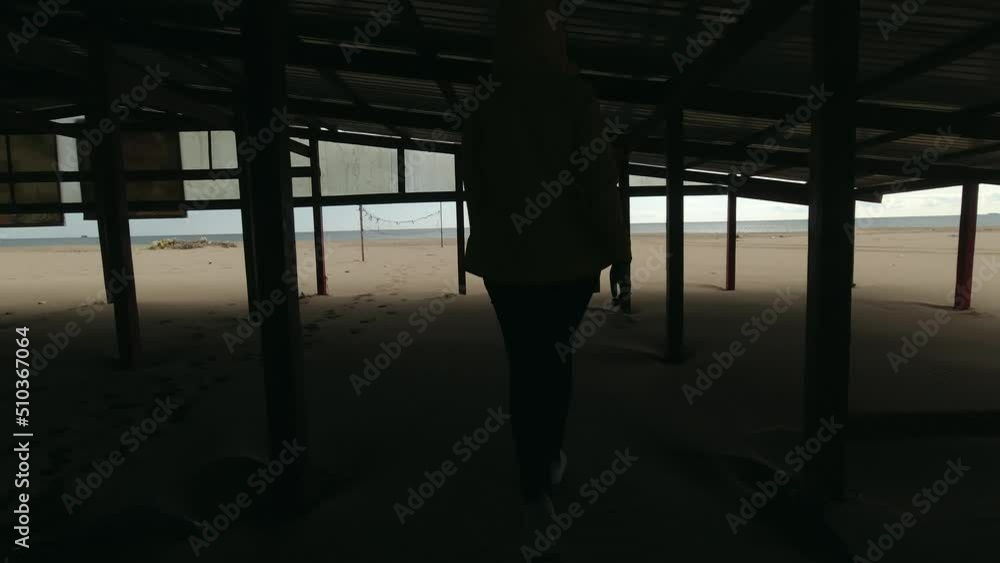 Young woman in yellow jacket walks along the sand to the sea under the abandoned construction Young woman in yellow jacket walks along the sand to the sea under the abandoned construction