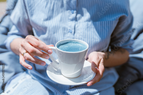 A girl in a blue shirt holds a cup of blue matcha tea with milk in her hands. Beautiful blue