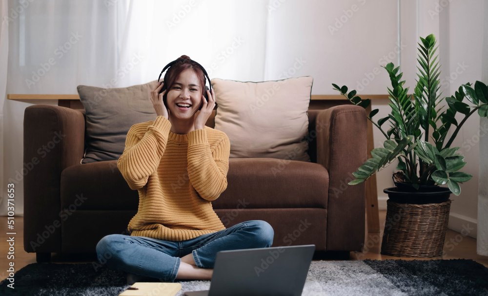 smiling girl looking at the camera Next to the laptop sit on the floor in front of the sofa and put on headphones to listen to music at home.