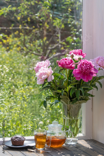 Delicious hot tea on the windowsill at home at summer day near garden and beautiful bouquet of flowers pink peonies  close up. Hot black tea in a glass teapot and cup