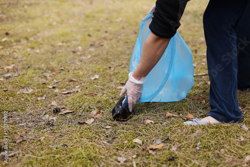 A woman hands in gloves collects and puts used plastic bottle into a blue trash bag. A volunteer cleans up the park on a sunny bright day. Clearing, pollution, ecology and plastic concept