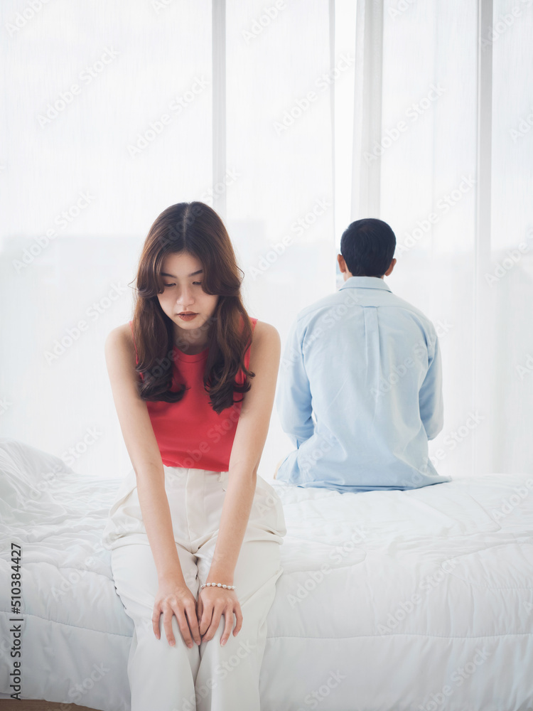The anxiety of Asian couple lovers on the bed. Sad young woman and man sitting on the bed with relationship difficulties feeling sad and thinking in the bedroom at home near the glass window. The anxiety of Asian couple lovers on the bed. Sad young woman and man sitting on the bed with relationship difficulties feeling sad and thinking in the bedroom at home near the glass window.