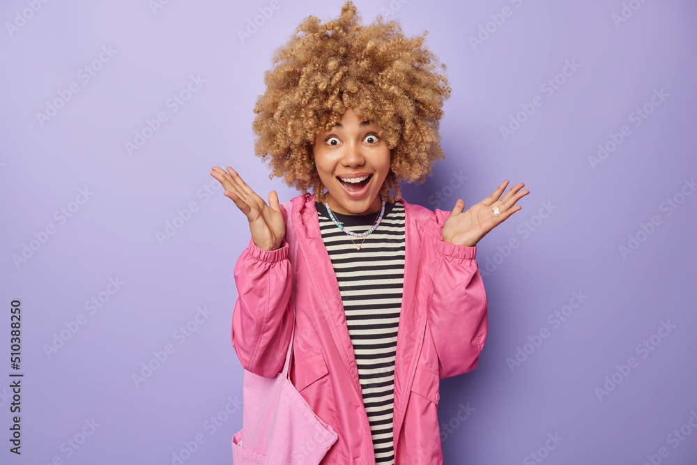 Happy amazed woman with curly hair spreads palms has surprised excitd expression wears casual striped jumper pink jacket and fabric bag isolated over purple background. Human reactions concept Happy amazed woman with curly hair spreads palms has surprised excitd expression wears casual striped jumper pink jacket and fabric bag isolated over purple background. Human reactions concept