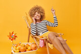 Cheerful woman has pleasant conversation via mobile phone points above shows something overhead has happy expression dressed in casual clothes poses on lounge chair against yellow background