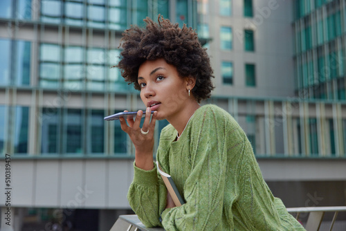 Horizontal shot of thoughtful curly haired woman uses cellphone for communication over speaker records voice message dressed casually poses against modern city building. Modern technologies.